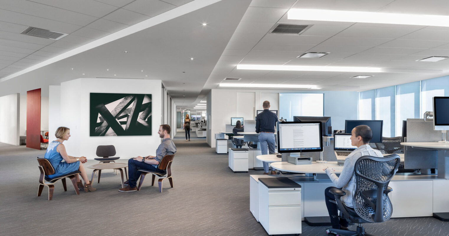 Open-plan office space with natural lighting, featuring modern furnishings—a mixture of ergonomic office chairs, personal workstations, and a casual seating area for discussions. White walls are complemented by a large, abstract green artwork, enhancing the contemporary feel.