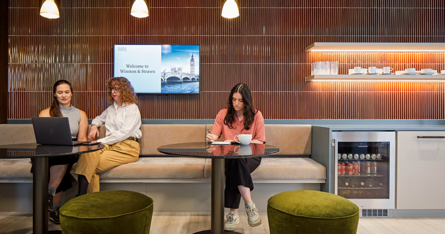 Modern office lounge with a warm wooden slat wall, complemented by sleek hanging pendant lights. A digital welcome sign adds a corporate flair, while plush green ottomans offer casual seating, beside a tidy kitchenette with a beverage fridge.