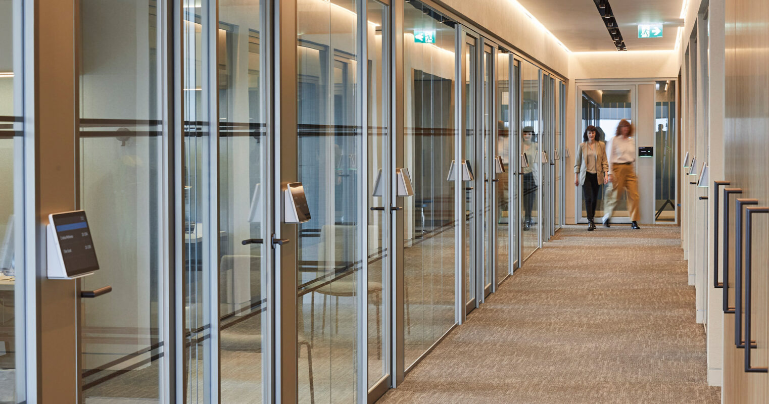 Modern office corridor featuring full-height glass partition walls, warm LED lighting, and sleek brushed metal door handles. A textured neutral carpet complements the minimalist design, enhancing the professional setting with people in motion, suggesting activity and collaboration.