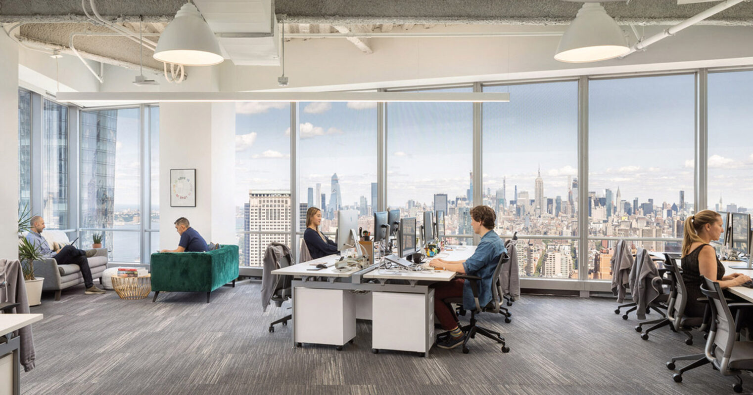 Open-plan office space with full-height windows offering a panoramic cityscape. Exposed concrete ceiling and pendant lighting complement the modern aesthetic, interspersed with ergonomic workstations and informal seating areas for collaborative work. Natural light floods the space, creating a vibrant and productive environment.