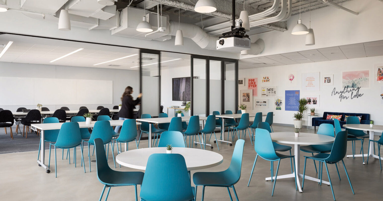 A modern office break room featuring exposed concrete ceilings with industrial pendant lighting. Turquoise chairs surround white, minimalist tables, creating a vibrant contrast against the neutral floor. Glass partitions delineate a meeting area, while motivational posters adorn the walls, promoting a creative work environment.