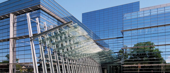 Modern glass building facade with a complex, transparent canopy structure supported by white steel beams, blending indoor and outdoor spaces under a clear blue sky.