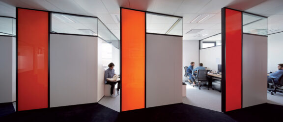 Modern office interior features glass partition walls with bold orange and white color blocking, enhancing the space's openness and fostering a collaborative yet private work environment. Individuals are engaged in tasks, illustrating the functionality of the design.