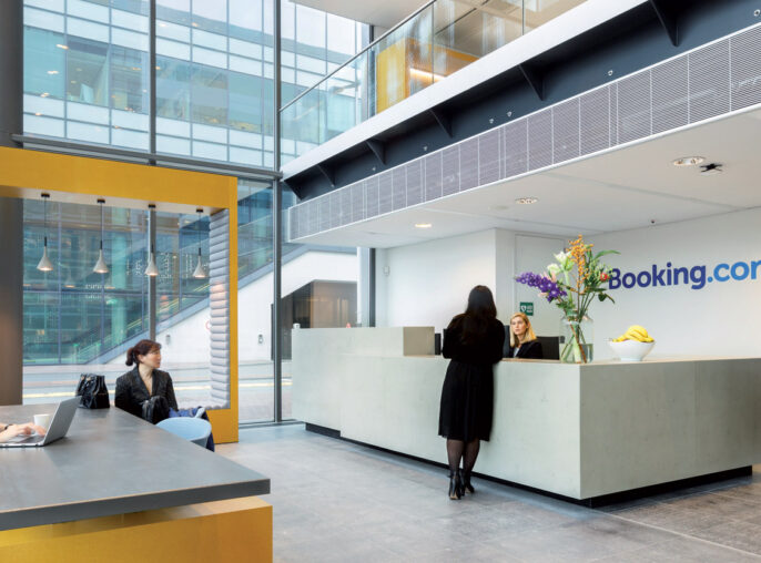 Modern office lobby with high ceilings and full-height windows, featuring a sleek white reception desk with a bold yellow accent panel. Two people work behind the counter, while others sit at a minimalist yellow bench, using laptops. The space is illuminated by abundant natural light.