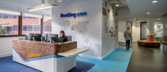 Contemporary office reception area featuring a world map mural on white tiled background. Wood-paneled reception desk with etched city map of Cambridge, against a vibrant blue carpet, enhancing the travel theme. Exposed ceiling pipes add an industrial touch contrasting with the welcoming ambiance.