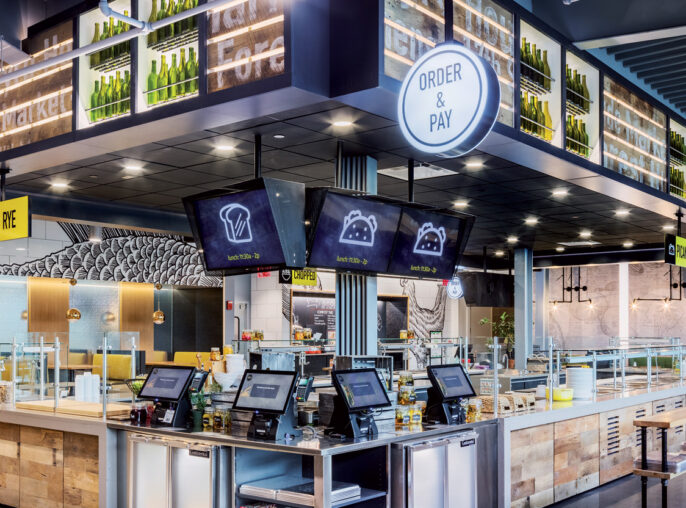 Modern fast-casual restaurant interior featuring an expansive order counter with digital menus, sleek black pendant lighting, and white subway tile backsplash. Organized seating zones and abundant use of natural wood tones complement the industrial chic vibe exemplified by exposed ductwork.