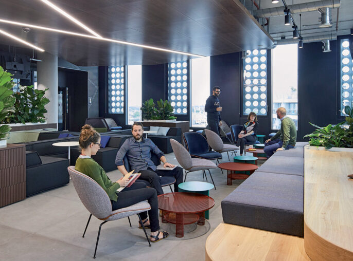 Spacious modern office break area featuring a mix of seating options, from plush sofas to wooden benches, under a distinctive lighting scheme with a dynamic, wavy LED track. A communal kitchen is anchored by potted plants, promoting an inviting and comfortable collaborative workspace.
