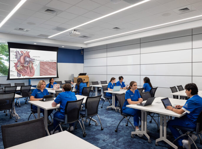 Spacious classroom featuring modern movable tables and chairs with a blue and white color scheme. The front wall displays a large projection screen, while ample natural light enhances the learning environment.