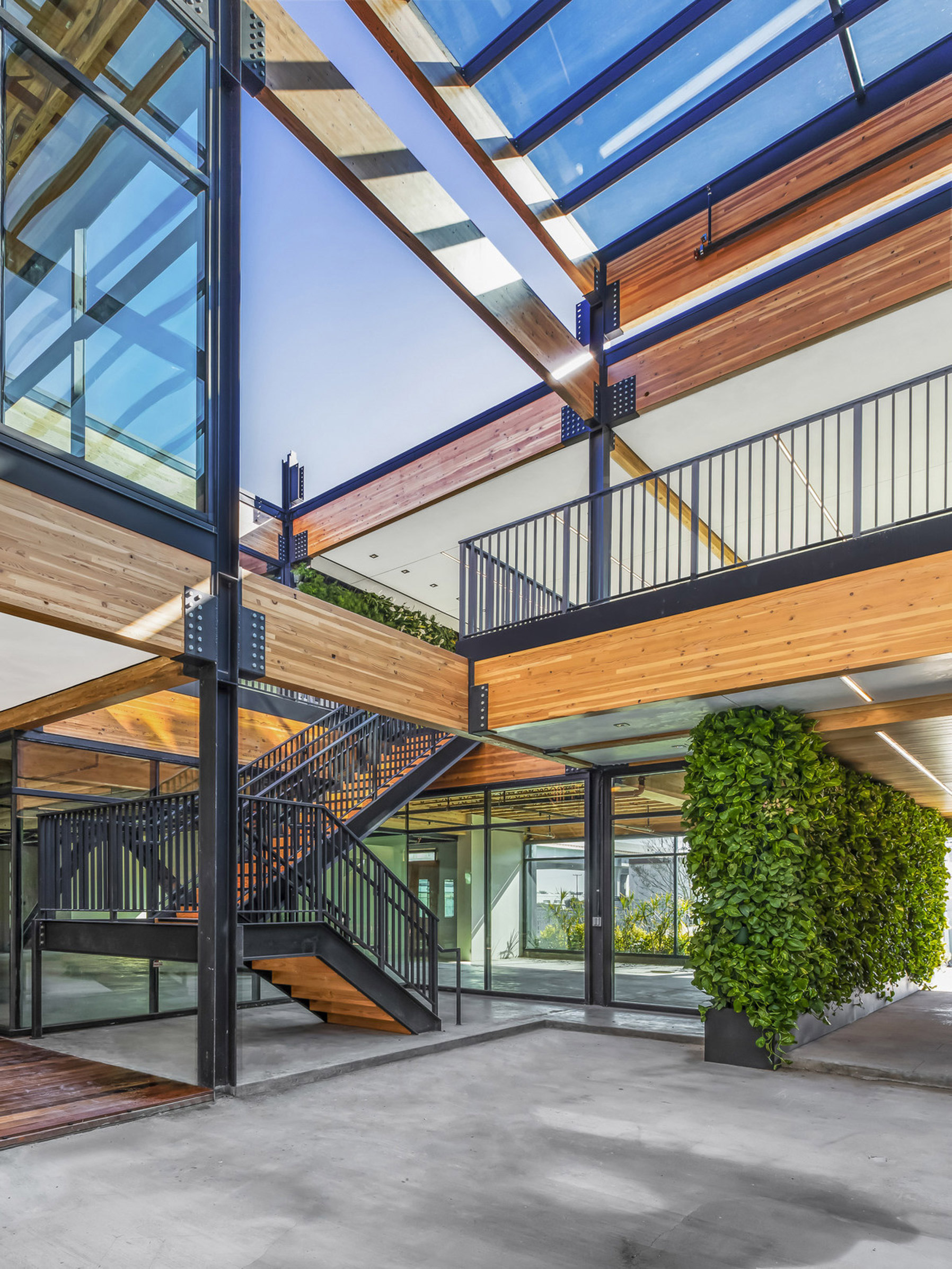 Modern open staircase with steel beams and wooden steps, flanked by a glass facade and skylights, leading to an upper-level gallery featuring natural wood flooring and railing, complemented by an indoor vertical garden.
