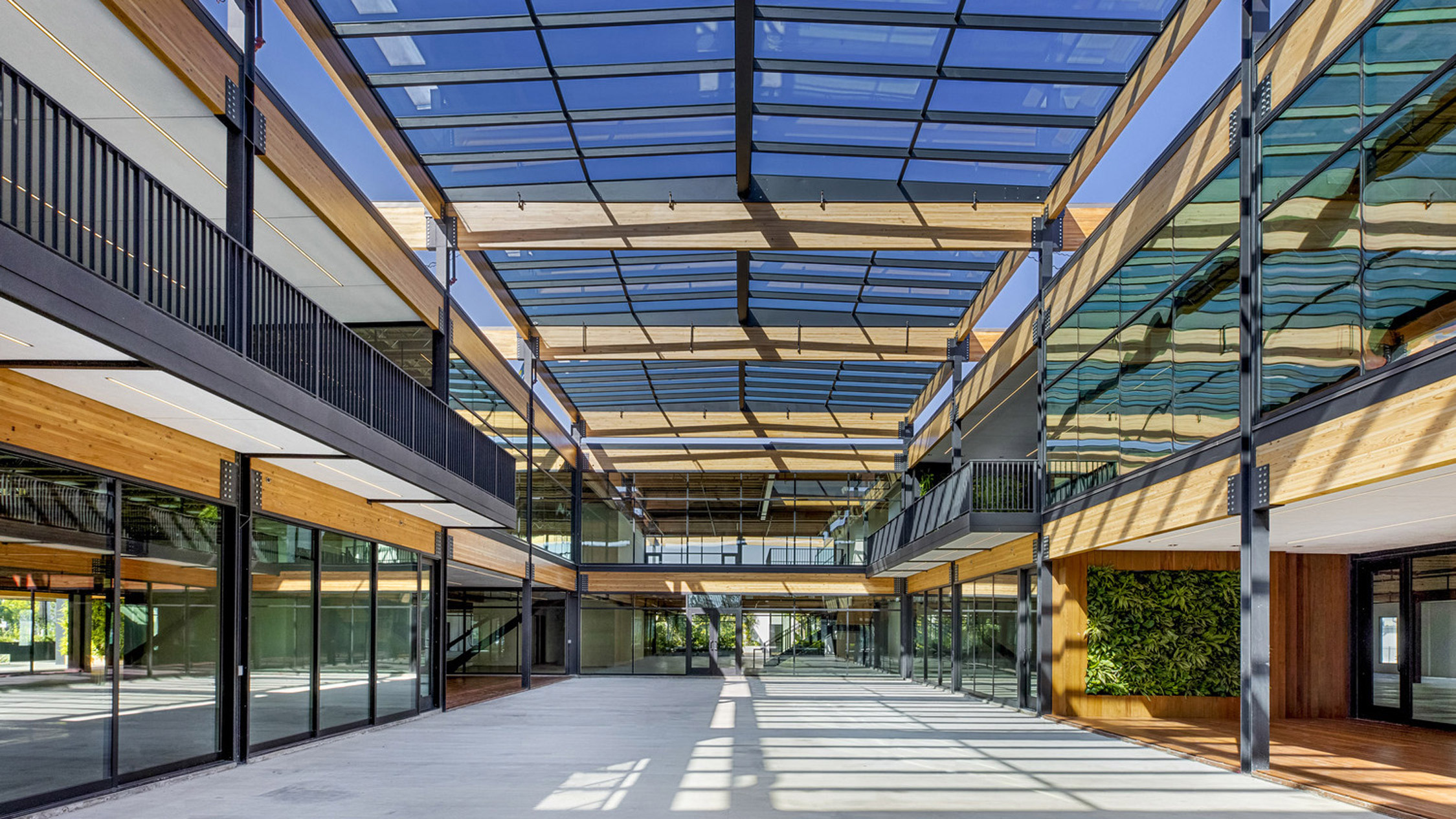 Spacious atrium with a vaulted glass ceiling allowing natural light to illuminate the sleek, wooden beams and modern, open-plan walkways flanking a central courtyard.