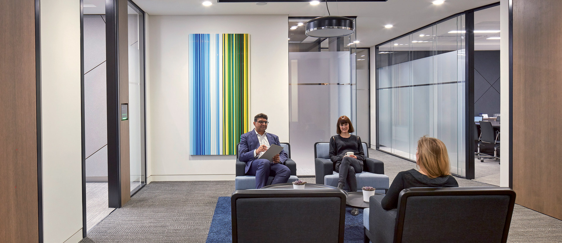 Modern office lounge space with sleek charcoal sofas, positioned around a low-profile coffee table. A vibrant, vertical stripe painting adds a pop of color against the neutral walls. Overhead, a circular pendant light fixture anchors the seating area, while floor-to-ceiling glass partitions provide transparency to adjoining workspaces.