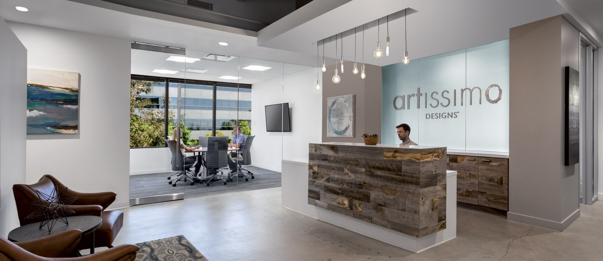 Modern office lobby with rustic wooden reception desk and sleek, white brand wall signage. Pendant lights hang above, contrasted by polished concrete floors and contemporary furniture, featuring expansive windows with outdoor patio access.