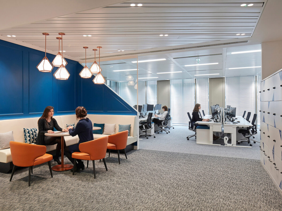 Modern office interior showcasing a harmonious blend of functional workspace and informal meeting area. Vibrant blue wall panels contrast with warm orange chairs, while sleek pendant lights add a contemporary touch above a cozy seating nook. Open-plan desks with ergonomic chairs promote a collaborative environment.