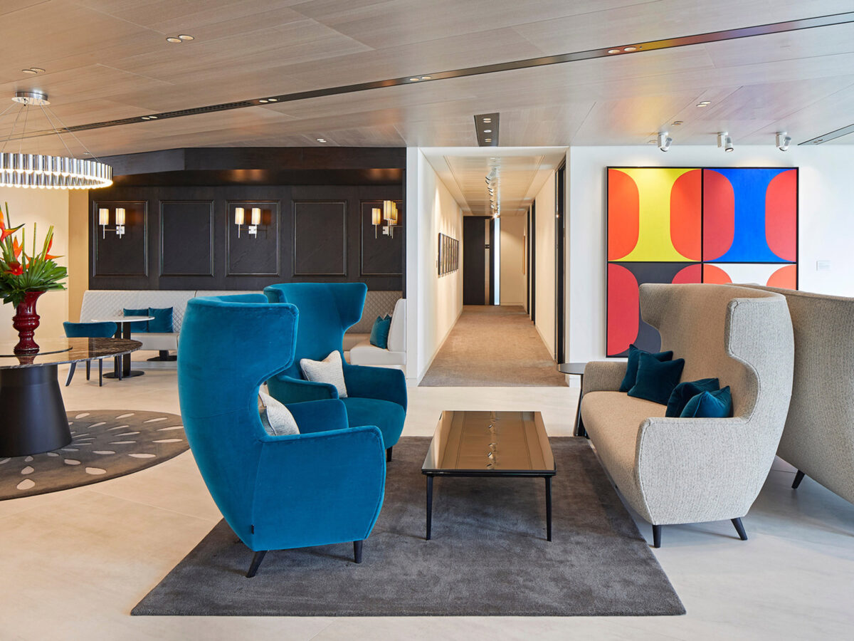 Spacious modern lounge area featuring teal armchairs, a sleek glass coffee table, and an abstract multicolored wall art. Earthy tones blend with the cool color palette under warm lighting, alongside dark wood panels and subtle geometric patterns in the carpet design.