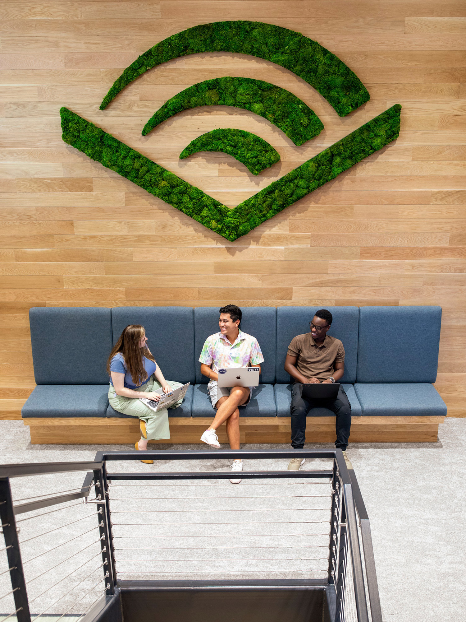 A vibrant workplace lounge with a wooden wall featuring a large green Wi-Fi symbol, flanked by a comfortable blue bench where three individuals engage in conversation and work.