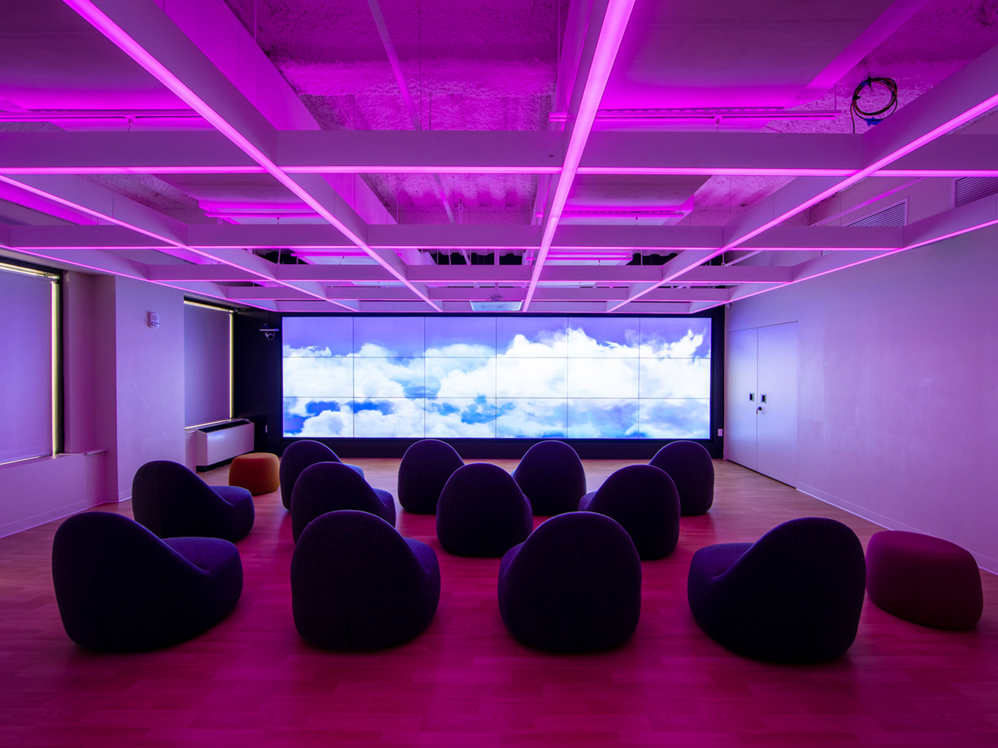 Modern interior with vibrant pink and purple LED lighting on the ceiling, complementing the dark beanbag chairs scattered across the carpeted floor, in front of a panoramic screen displaying a serene sky.