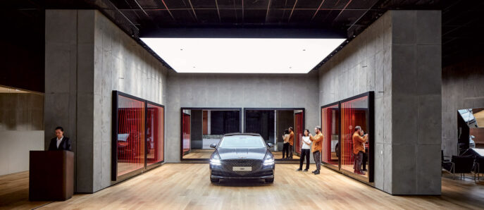 Sleek automotive showroom interior featuring exposed concrete walls and a polished floor reflecting a luxury car centerpiece. Red-framed glass doors add a vibrant contrast, while strategic lighting accentuates the modern, minimalist design. Staff and potential customers populate the space, enhancing the upscale atmosphere.