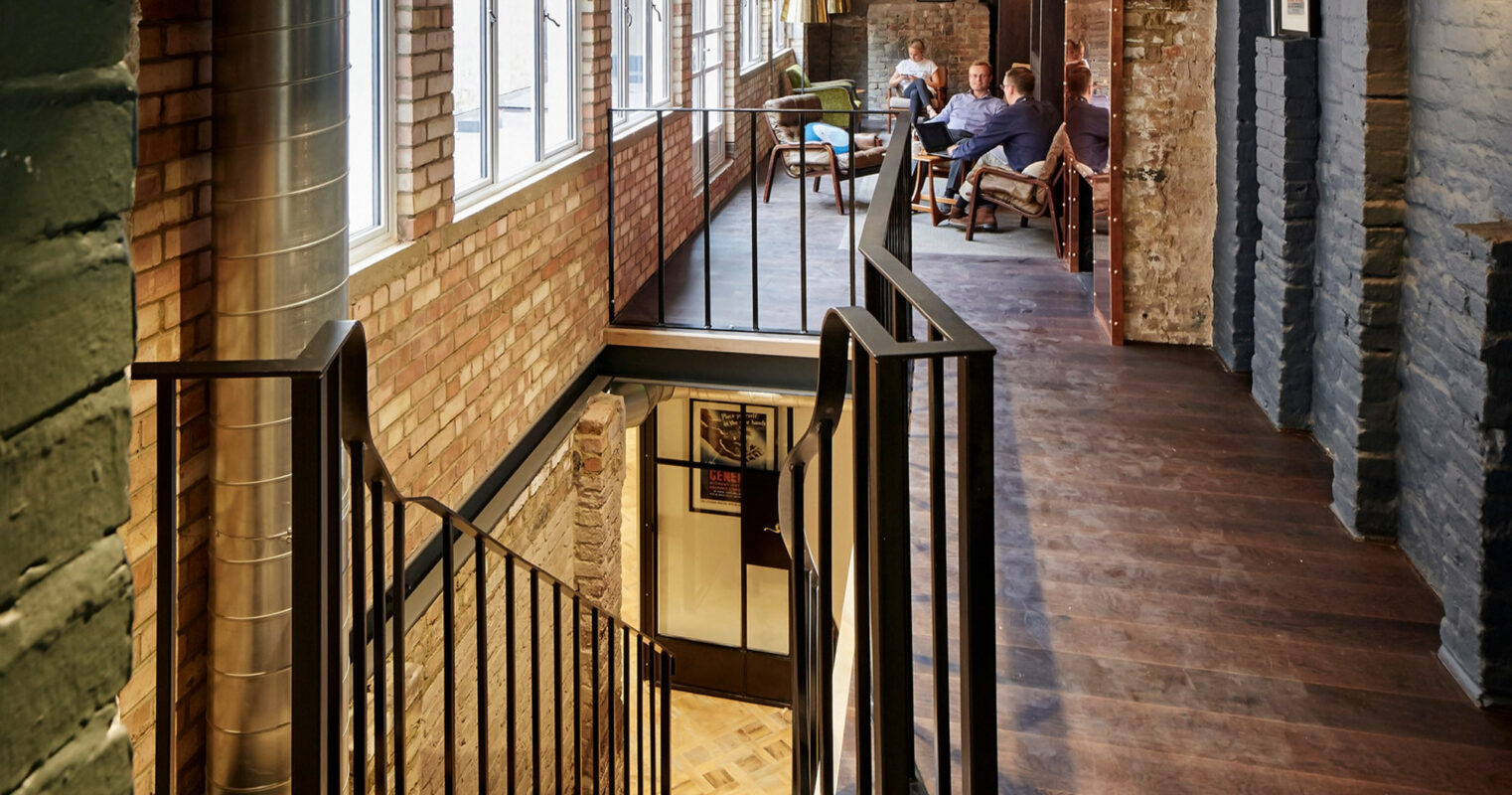 Stairway in a multi-level office space with brick and slate accents, iron railings, and vintage lighting, leading to an informal meeting area, blending industrial design with warmth and hospitality.