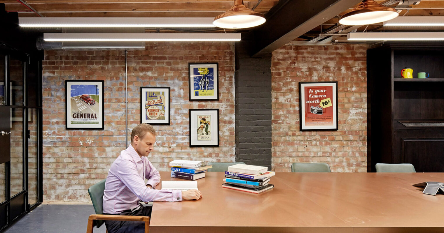 Brick-walled workspace with a long wooden table and eclectic wall art, illuminated by linear lighting, providing a creative and informal meeting environment.