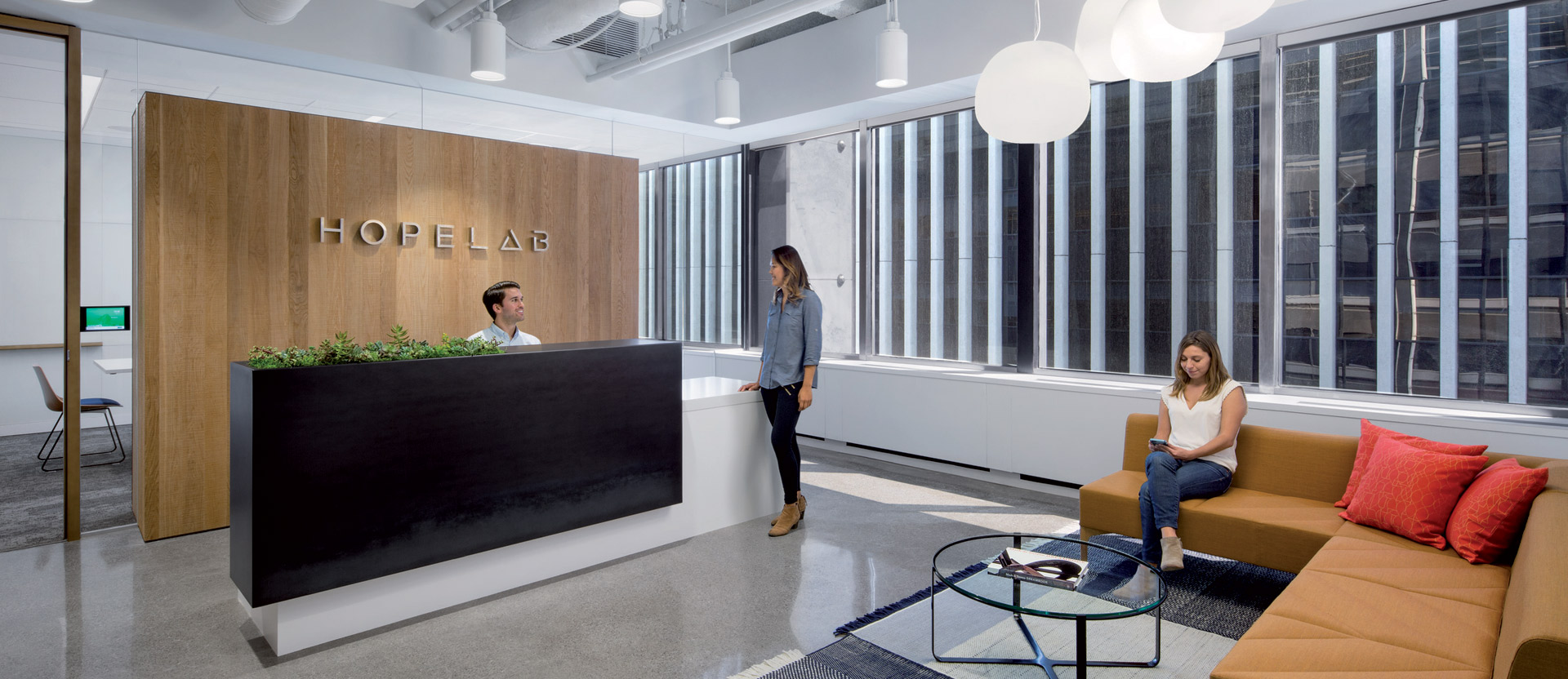 Spacious office reception area featuring a contemporary design with clean lines, an exposed ceiling, and modern pendant lighting. A wooden panel with the company's name complements the industrial background while greenery adds a natural touch. Comfortable seating arrangements invite casual interactions among the individuals present.
