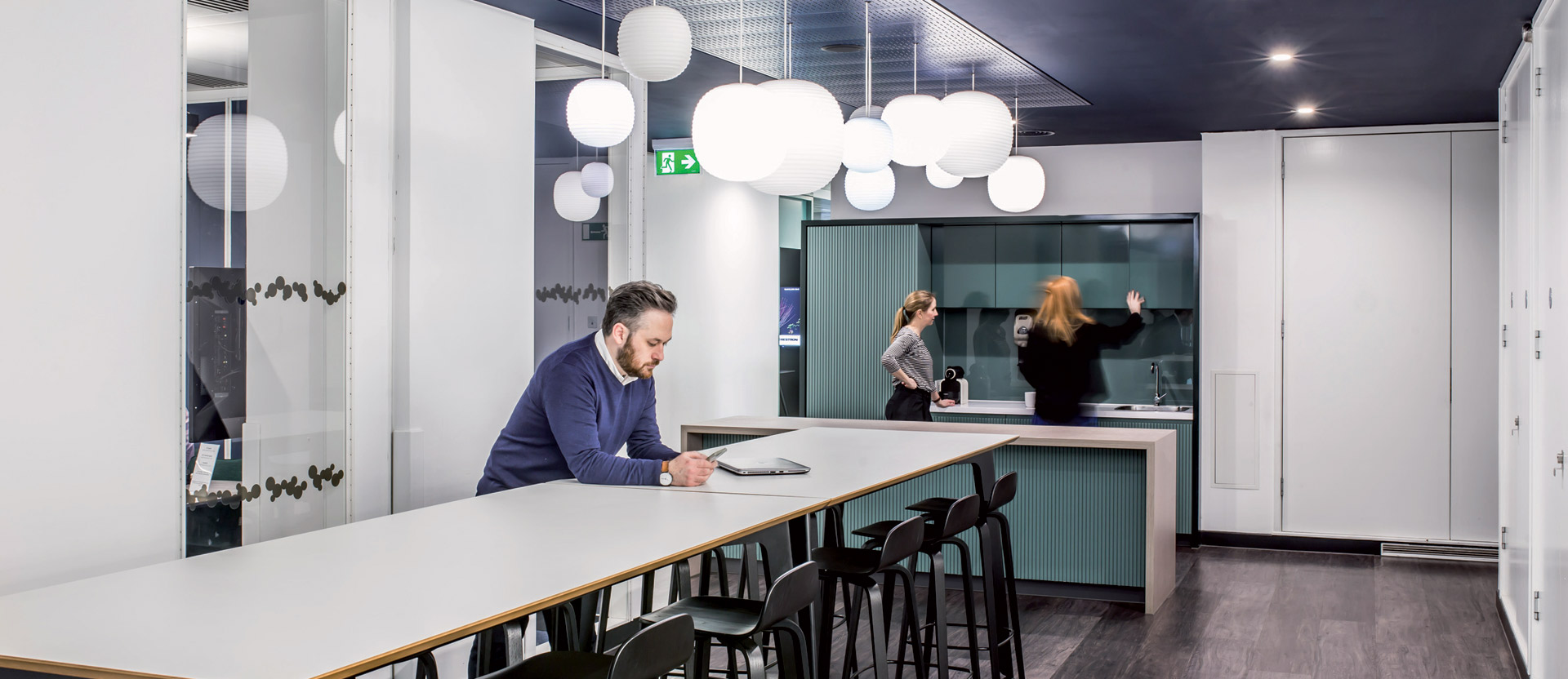 Modern office break room featuring teal cabinetry, sleek black countertops, and a variety of globe pendant lights suspended at varying heights. Professionals engage in work and conversation, emphasizing a functional yet stylish workspace designed for interaction and comfort.