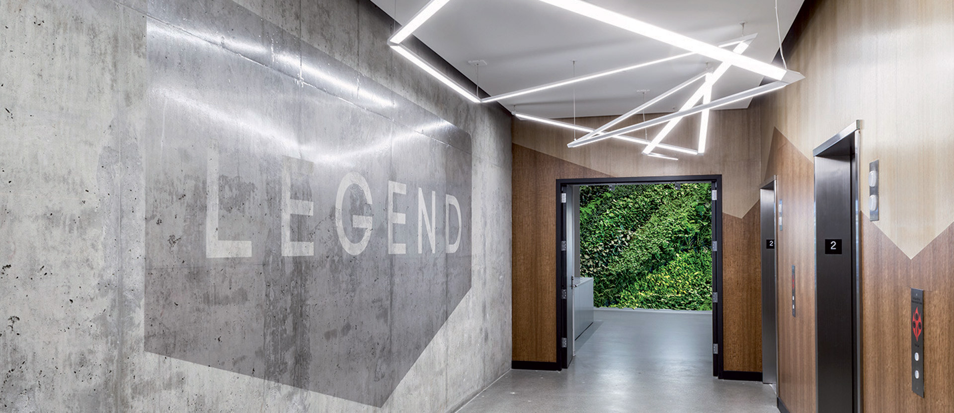 Modern hallway with geometric LED ceiling lights and polished concrete walls featuring a bold stenciled "LEGEND". Warm wooden doors contrast with the industrial aesthetics, complemented by a lush living green wall at the end of the corridor.