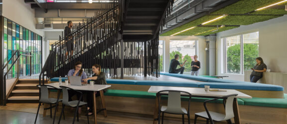 Contemporary open-plan office space featuring a mix of industrial and biophilic design elements with exposed ceiling pipes, cascading greenery, and central tiered seating area fostering collaboration.