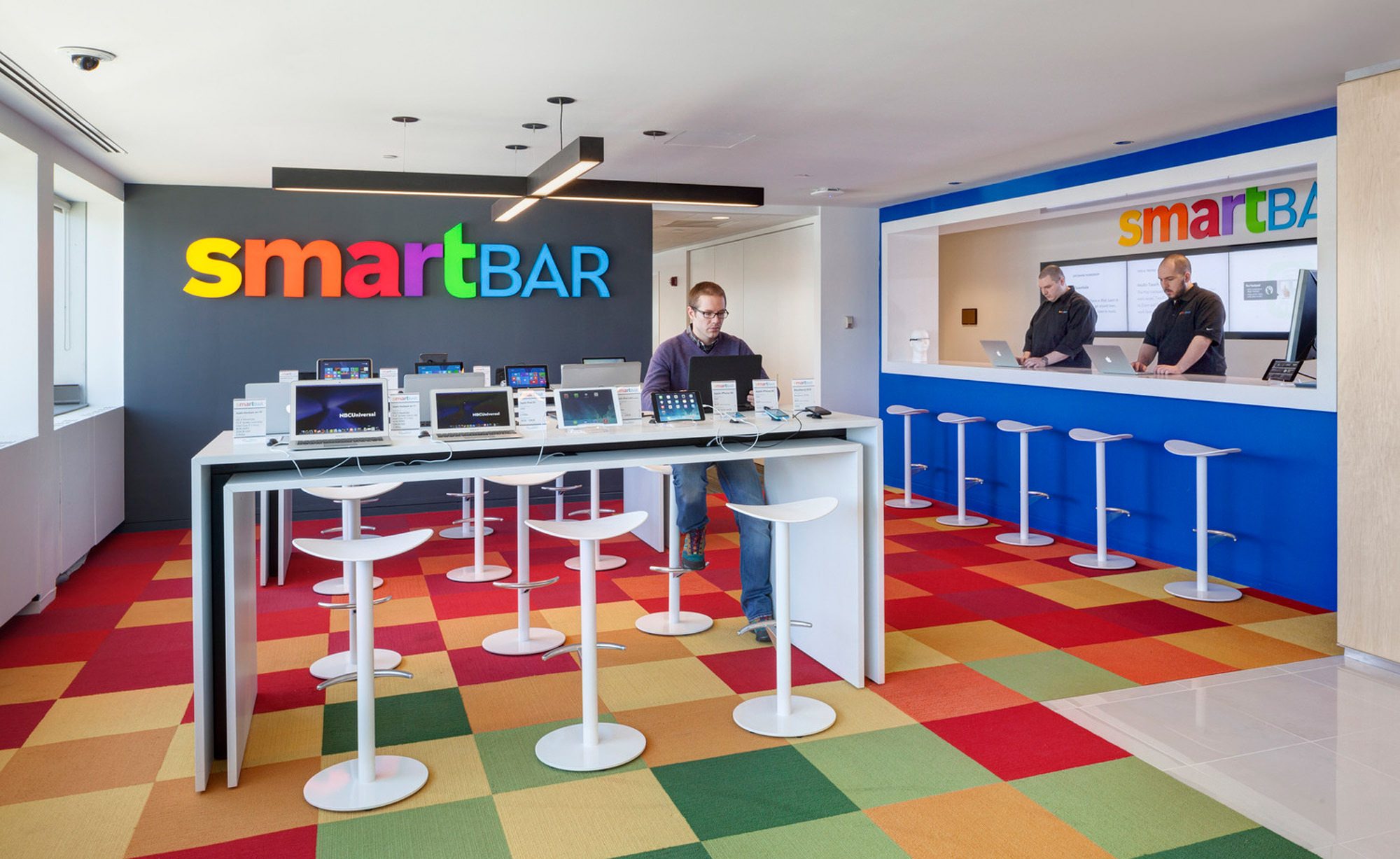 Modern office break area featuring a vibrant multicolored geometric-patterned carpet, white standing tables with bar stools, and minimalistic decor against an accent wall with 'smartBAR' branding. The space is designed for collaboration with an informal, energetic atmosphere.