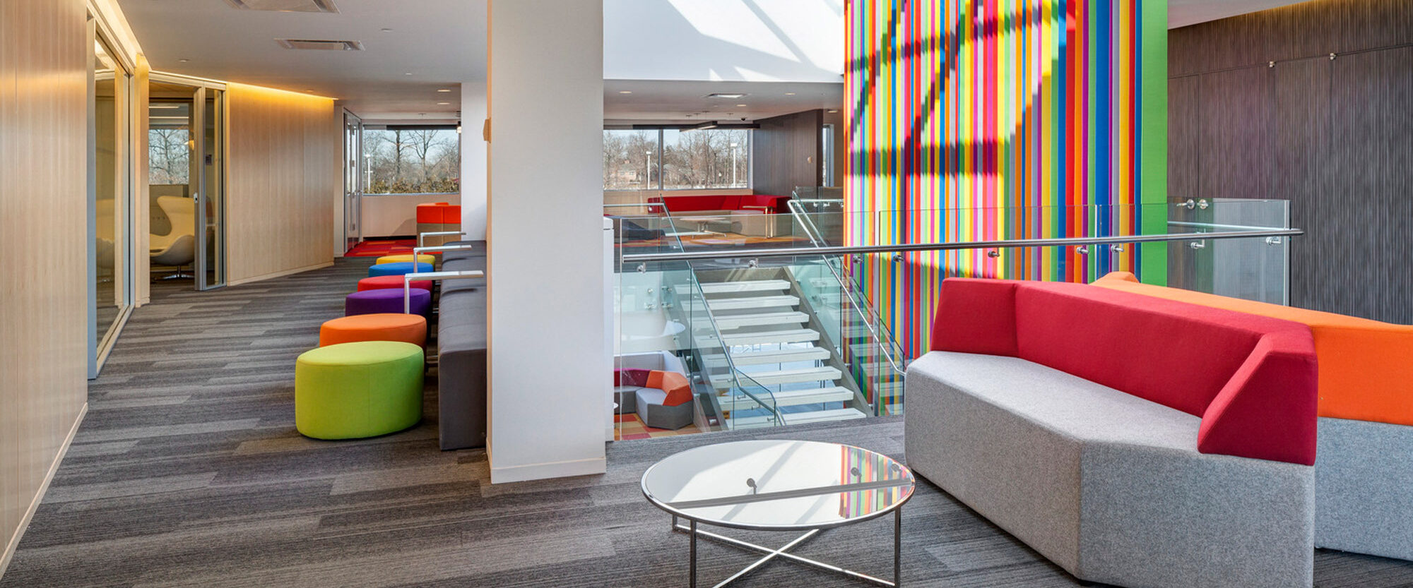 Bright modern office lobby featuring a colorful striped partition, gray carpeted flooring, minimalist glass stair railing, vibrant furniture accents, and ample natural lighting from skylights above.