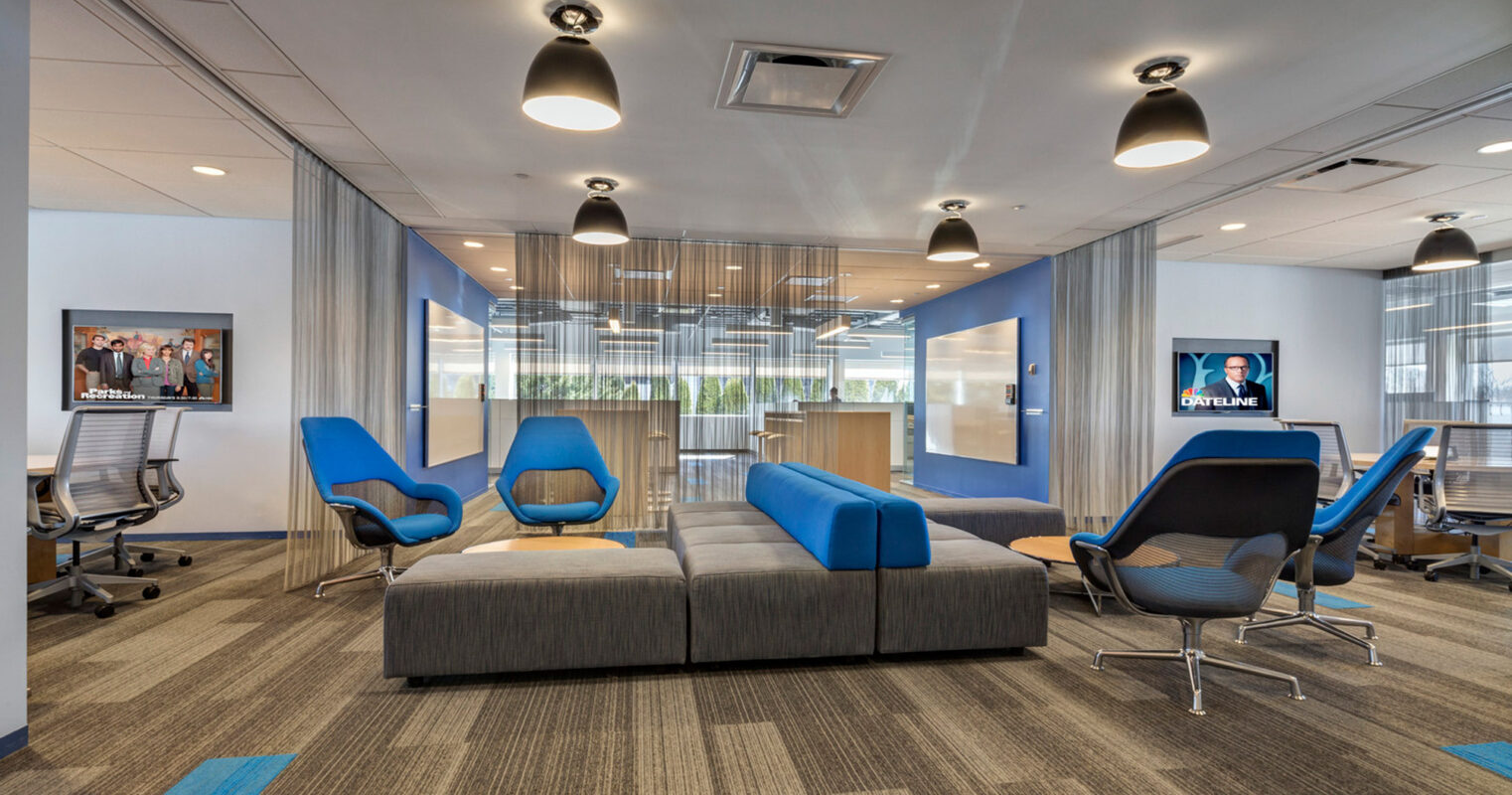 Open-plan office space featuring sleek, modular seating in shades of blue and gray, complemented by floor-to-ceiling windows with sheer drapery. Overhead, industrial-chic pendant lights illuminate the area. A trio of ergonomic office chairs and a multimedia display punctuate the room's functionality.