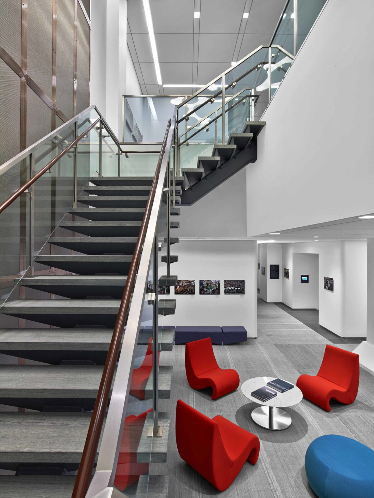 A spacious, modern lobby featuring a floating staircase with glass balustrades and wood accents. Below, vibrant red armchairs and blue ottomans provide a pop of color against the neutral palette of grey walls and white flooring. Artwork is strategically placed along the corridor for visual appeal.