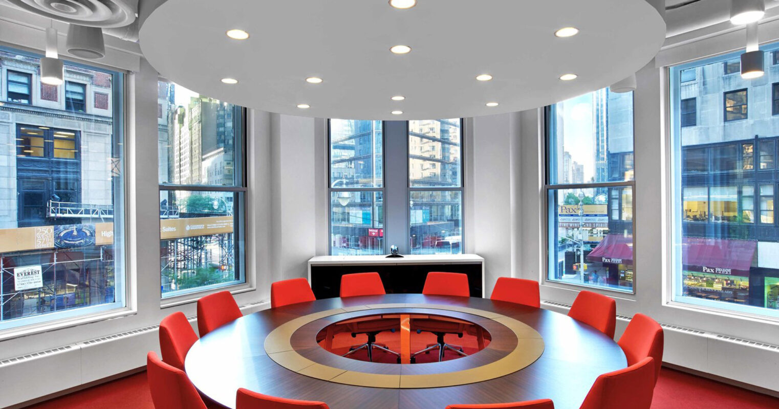 Bright conference room with large circular table surrounded by red chairs, a circular white pendant light above, and floor-to-ceiling windows offering a city view. The interior showcases a modern design with a vibrant color contrast.