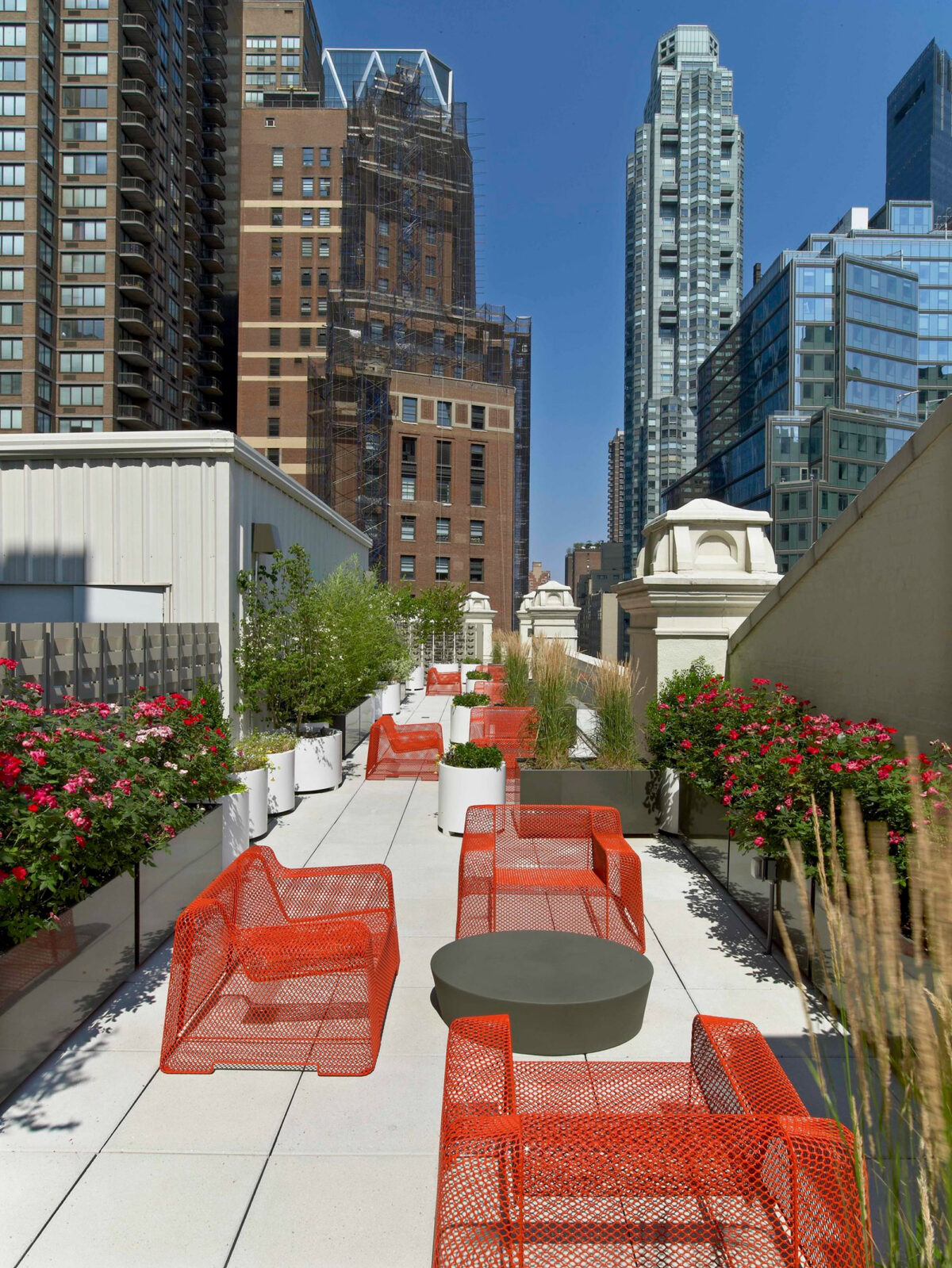Modern rooftop terrace featuring vibrant orange wire-frame chairs and cement coffee tables, complemented by lush greenery and urban backdrop with skyscrapers.