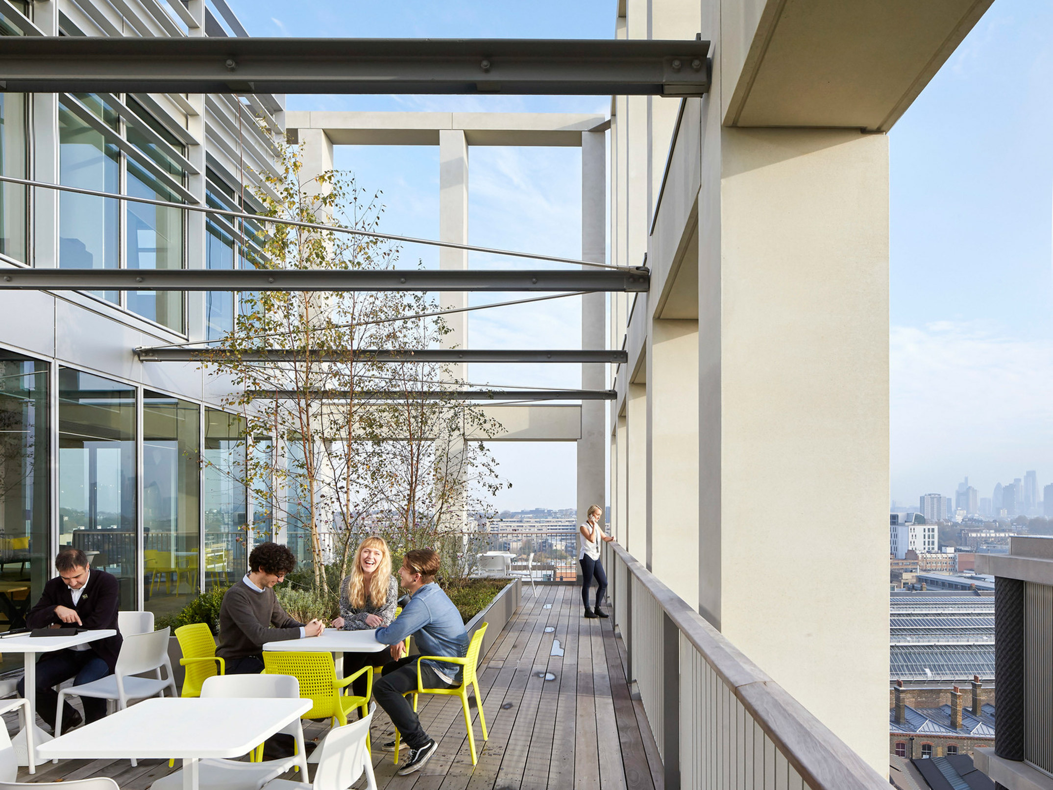 Contemporary urban balcony featuring glass railing, steel supports, and minimalist furniture with pops of color. Lush greenery accents the space, overlooking a cityscape under a clear sky. People engage in relaxed conversation, embodying a seamless blend of natural elements in a modern setting.