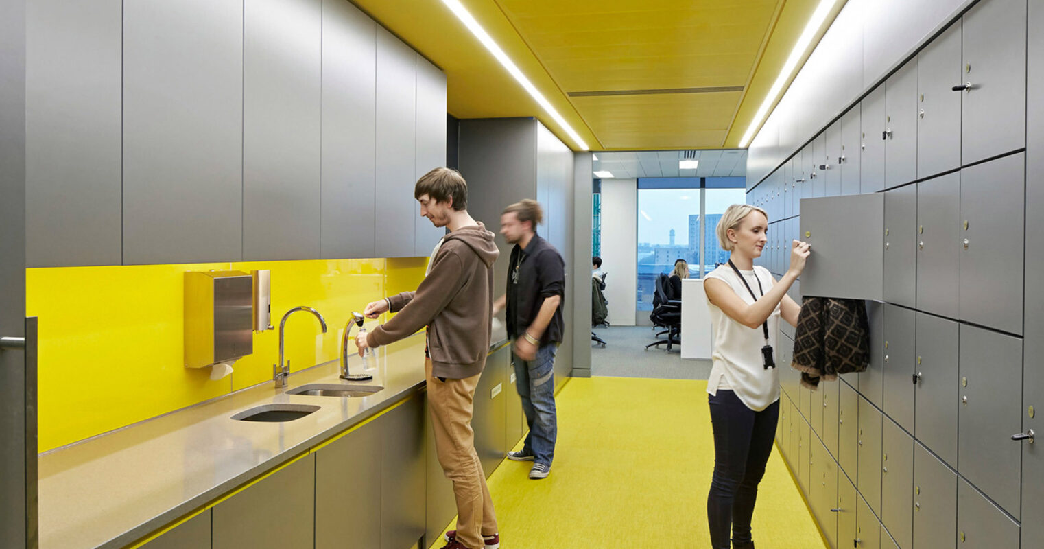 Modern office break room featuring lemon-yellow accents in backsplash and flooring, with sleek gray cabinetry and stainless steel fixtures. Natural light permeates the space, enhancing the vibrant color scheme and clean lines. Individuals engage casually, adding a dynamic element to the functional design.