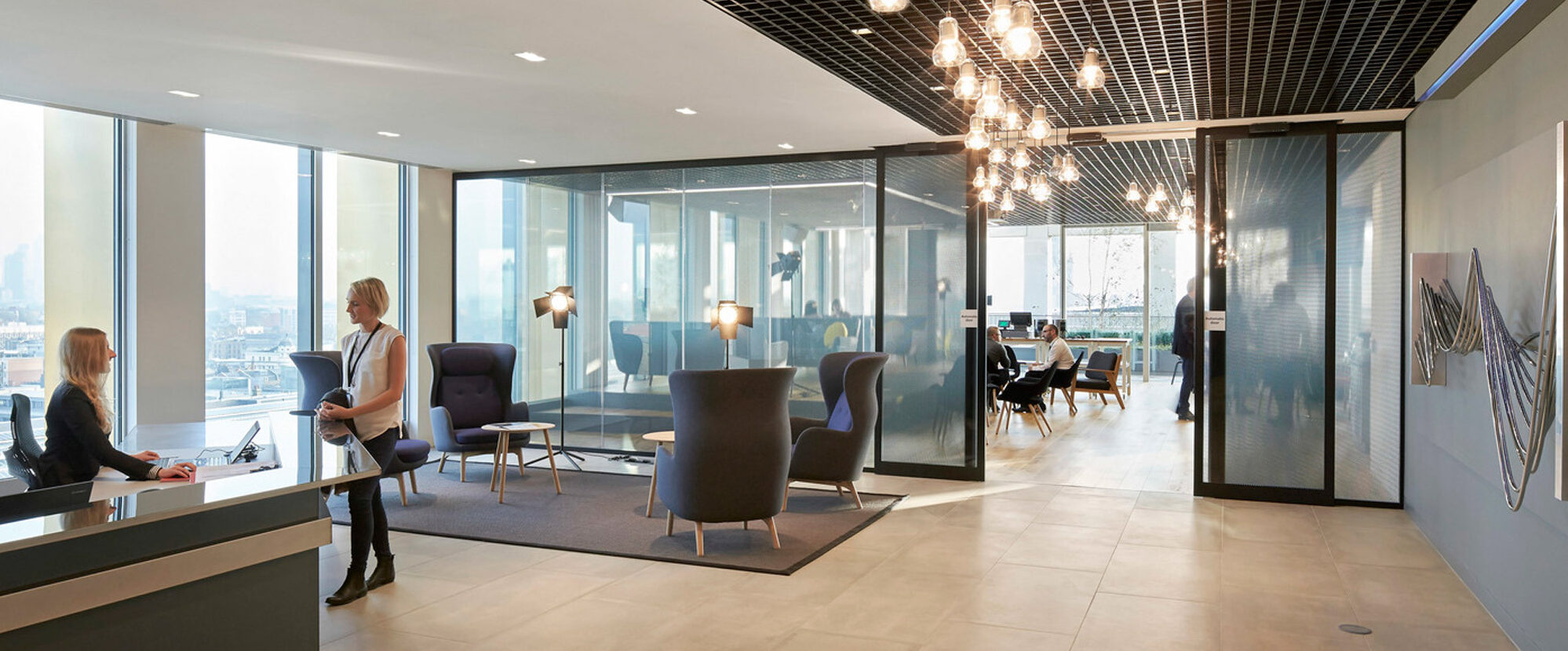 Modern office space showcasing an open layout with expansive windows offering cityscape views. A variety of blue armchairs and a sleek reception desk establish a professional, welcoming ambiance, complemented by unique geometric ceiling lights and discreet glass partitions.