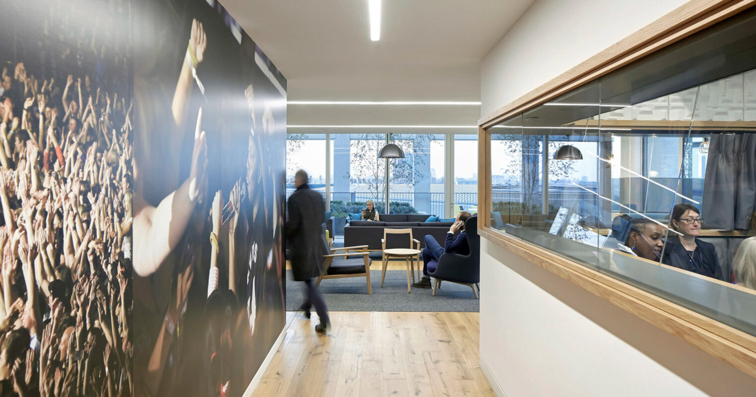 Modern office lobby featuring hardwood floors, minimalist furniture, a large photo mural wall with a crowd scene, sleek horizontal lighting, and a glimpse of an adjoining workspace through interior windows.
