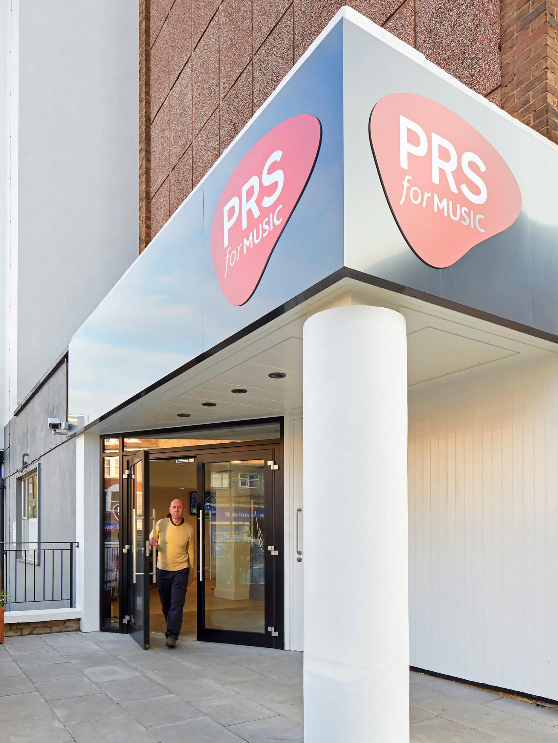Modern storefront with a bold PRS for Music sign in vibrant pink and blue, showcasing a clean, minimalist design with large glass doors, a sleek overhang, and an inviting, open entryway.