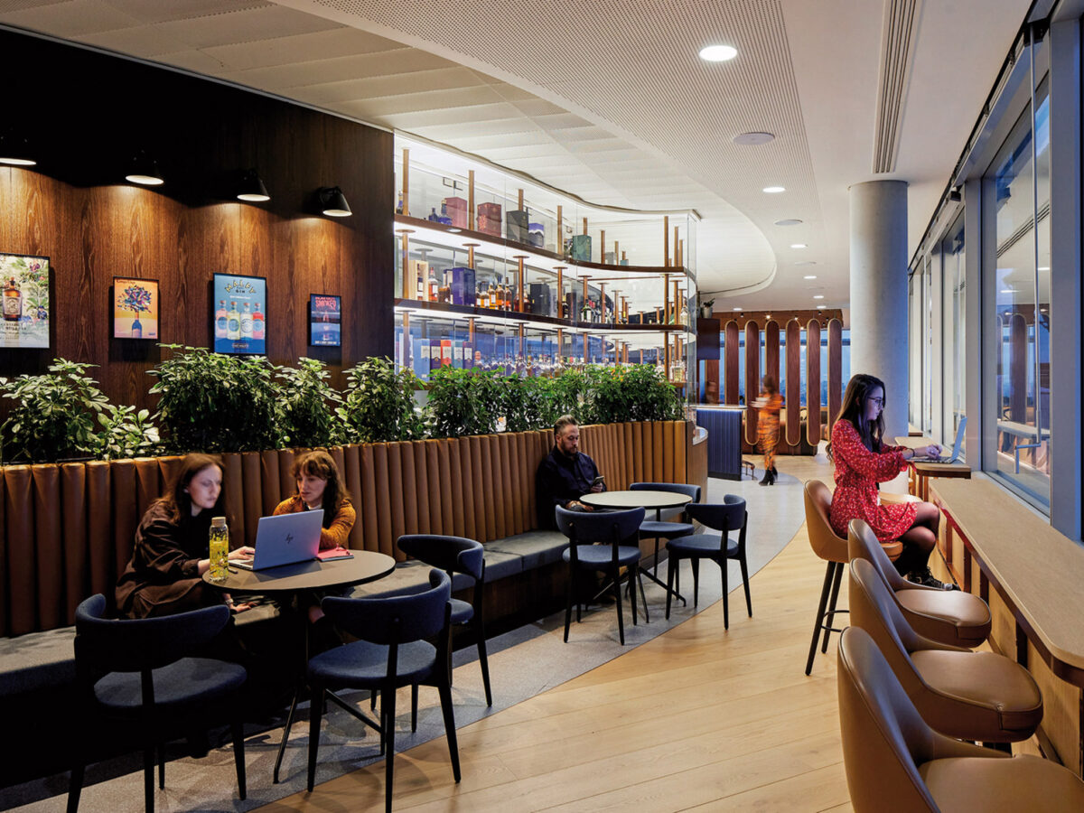 Modern café interior featuring a warm wood-panelled wall with shelving displaying books and décor. A series of upholstered booth seating provides privacy, while individuals interact or work independently amidst the ambient lighting and expansive windows offering ample natural light.