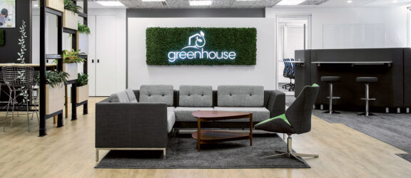 Modern office lobby featuring a neutral palette with grey sofas, a central wooden coffee table, and a moss wall displaying the "greenhouse" logo. Strategically placed greenery complements the natural aesthetic, while subtle lighting accentuates the space's clean lines and contemporary design.