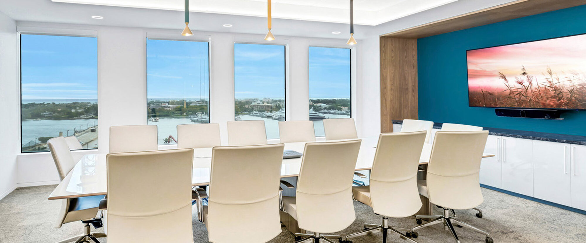 Modern conference room featuring an elongated white table with beige chairs, a wide-screen monitor on a blue accent wall, and pendant lighting in muted tones against a panoramic window backdrop.
