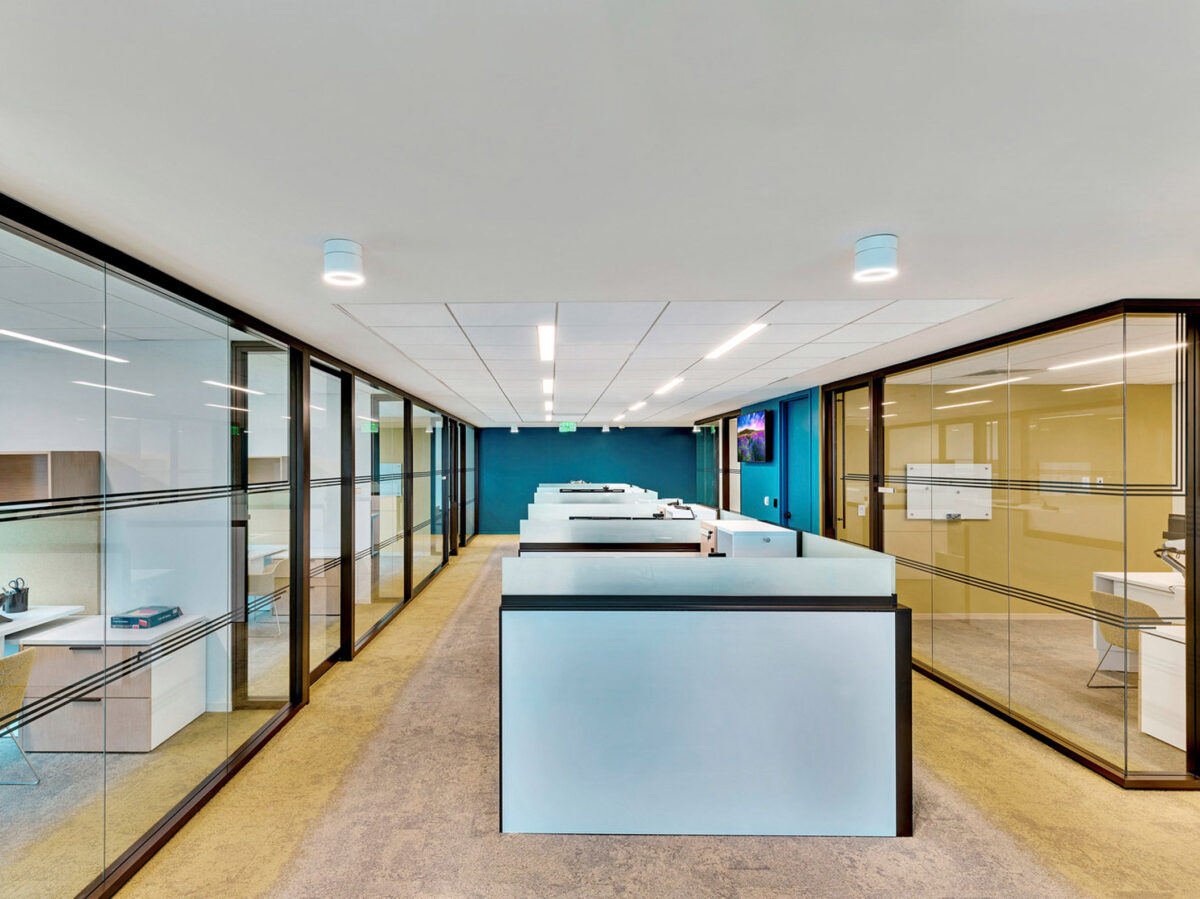 Modern office interior with glass partition walls, sleek white desks in a row, and overhead cylindrical lighting. The design emphasizes transparency and collaborative workspaces, accented by a vibrant blue carpet and abstract wall art.