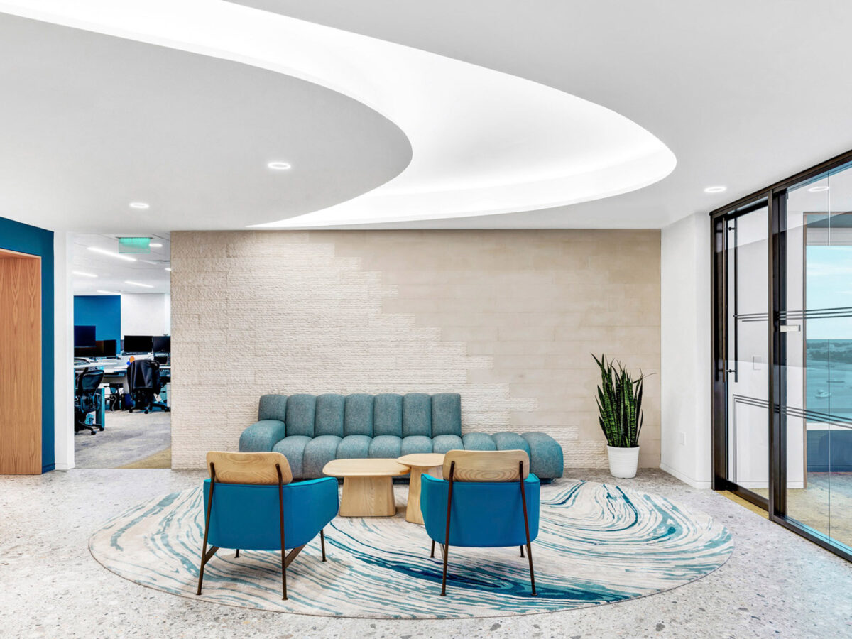 Elegant modern office lounge with a sculptural white ceiling feature, teal and blue armchairs gathered on an abstract-patterned rug, and a textured beige wall, complemented by natural light streaming in from adjacent glass doors.