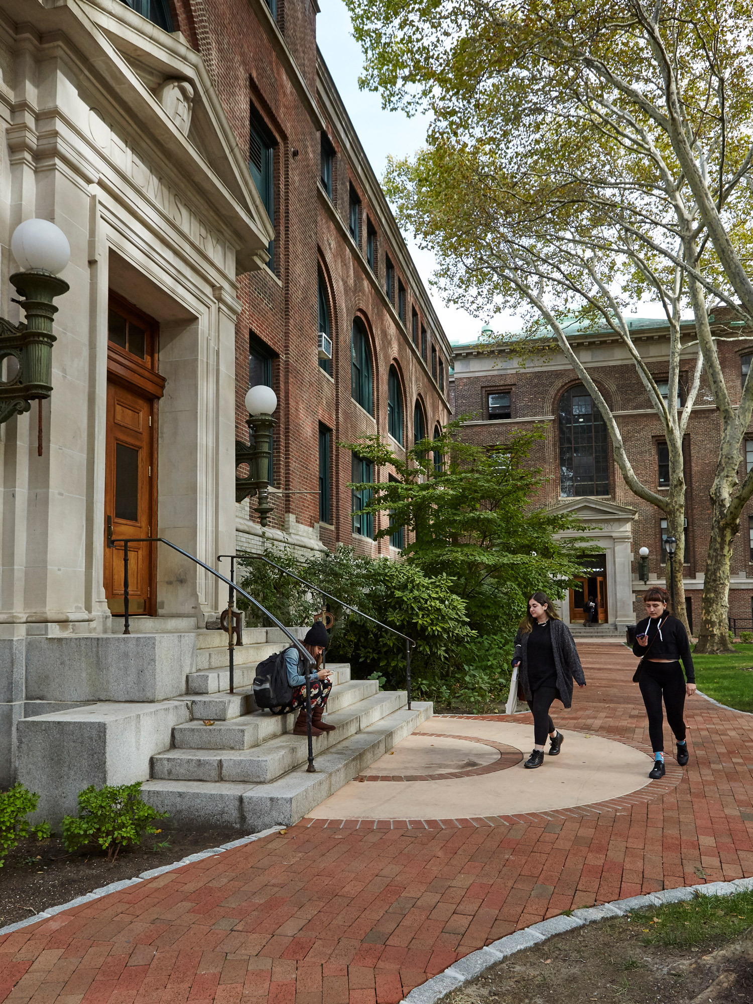 Elegant collegiate architecture with a red brick facade, grand white-trimmed windows, and a stately entrance featuring a wooden door flanked by vintage lanterns, set against a backdrop of verdant trees and a manicured lawn, with students traversing the serene campus setting.