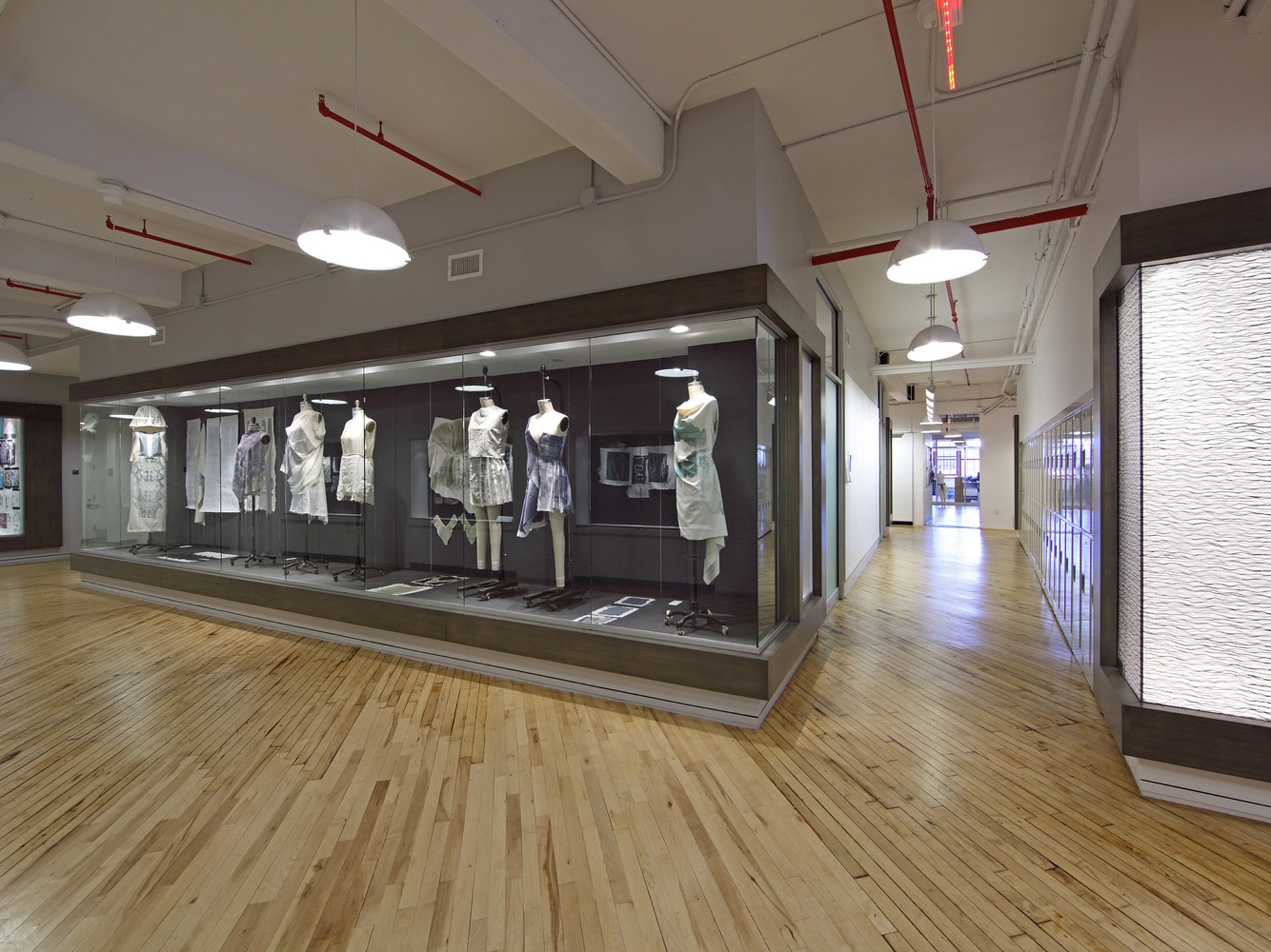 Modern gallery space featuring a display case with historic clothing, polished wooden floors, white walls, and industrial ceiling with exposed red ductwork. LED track lighting accentuates the exhibit, blending function with contemporary design.