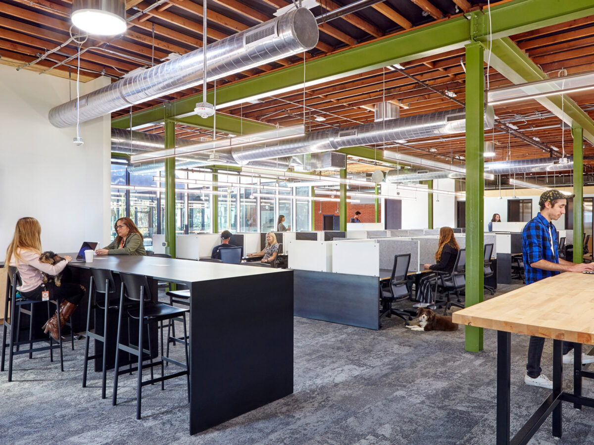 Modern open-plan office with exposed wooden beams and HVAC ductwork. Sleek, black modular workstations are paired with ergonomic chairs. Natural light streams in, complementing the vibrant green structural columns and creating a lively workspace that fosters collaboration and innovation.