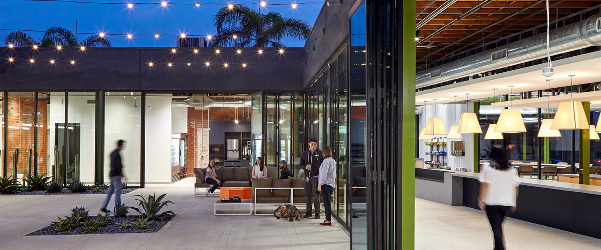Open-plan office space seamlessly integrating indoor and outdoor areas with retractable glass walls, exposed ceiling beams, and pendant lighting. The design incorporates a modern industrial look with green plants adding a touch of nature.
