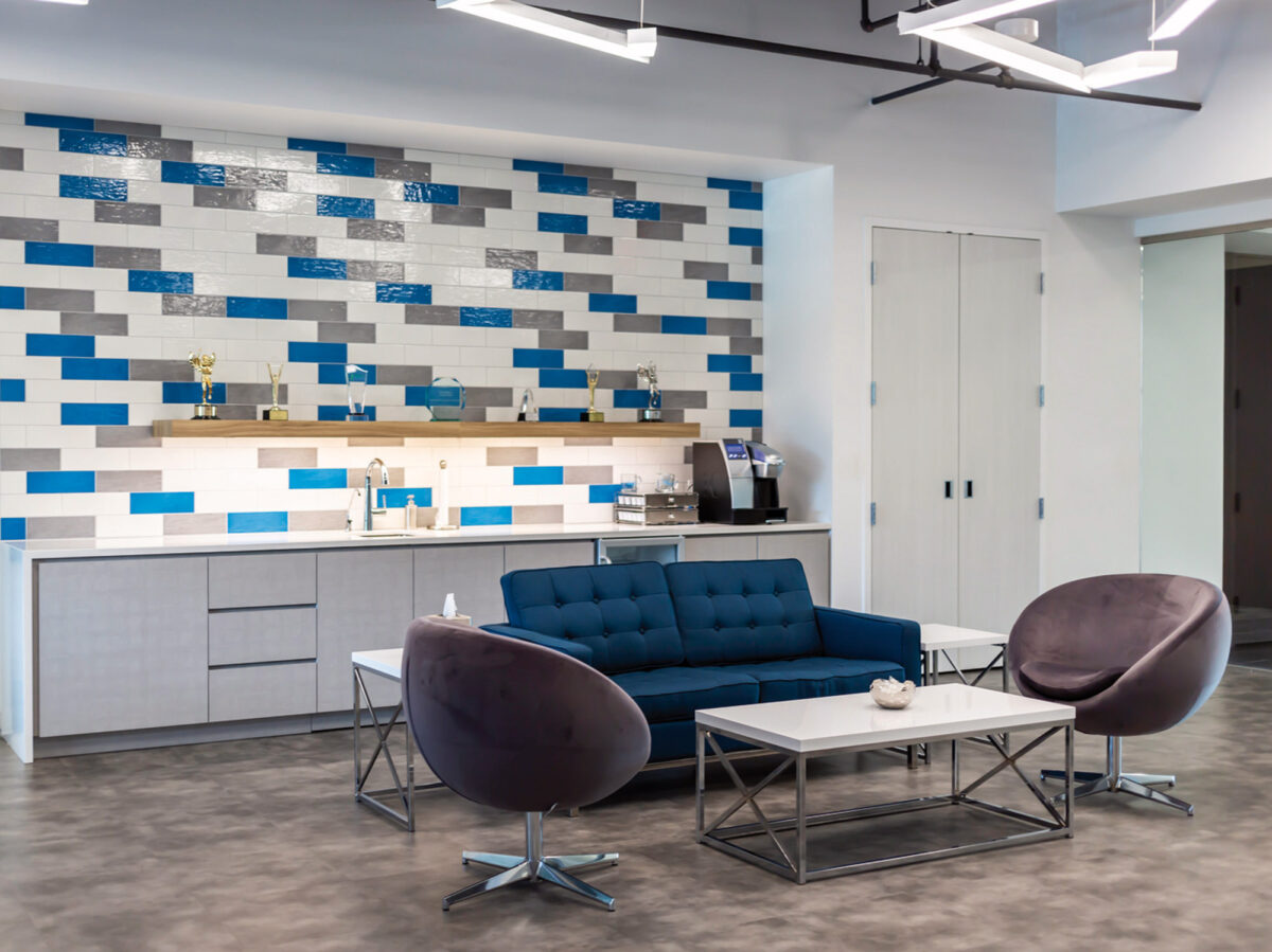 Modern office lounge featuring a deep blue tufted sofa, complemented by two eggplant hued swivel chairs, and a sleek white coffee table. A geometric wall pattern with blue accent tiles creates a vibrant backdrop, complementing the polished concrete floor.