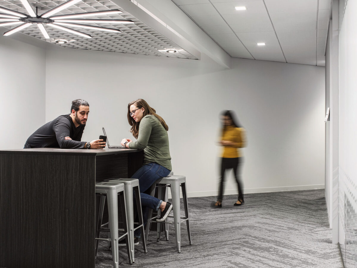 Modern office break area with minimalist design, featuring a sleek black counter, gray stools, geometric ceiling panels, and ambient lighting. Two individuals engaged in conversation while using laptops, with another person walking by in the background, illustrating a dynamic work environment.