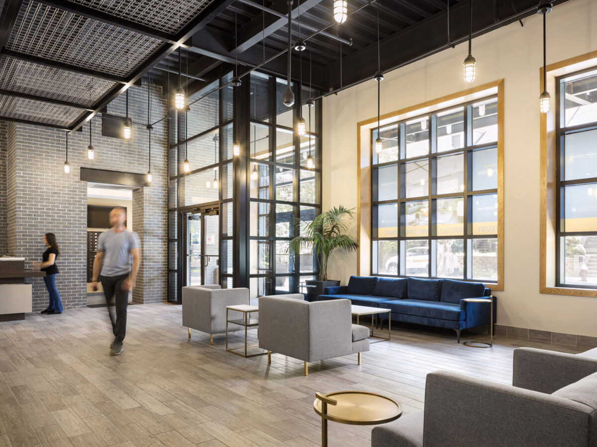 Modern industrial-style lobby featuring high ceilings with exposed ductwork, large arched windows allowing ample natural light, a sleek blue velvet sofa, accent armchairs, and brass-toned circular coffee tables. A reception desk and busy professionals add to the functional yet stylish space.